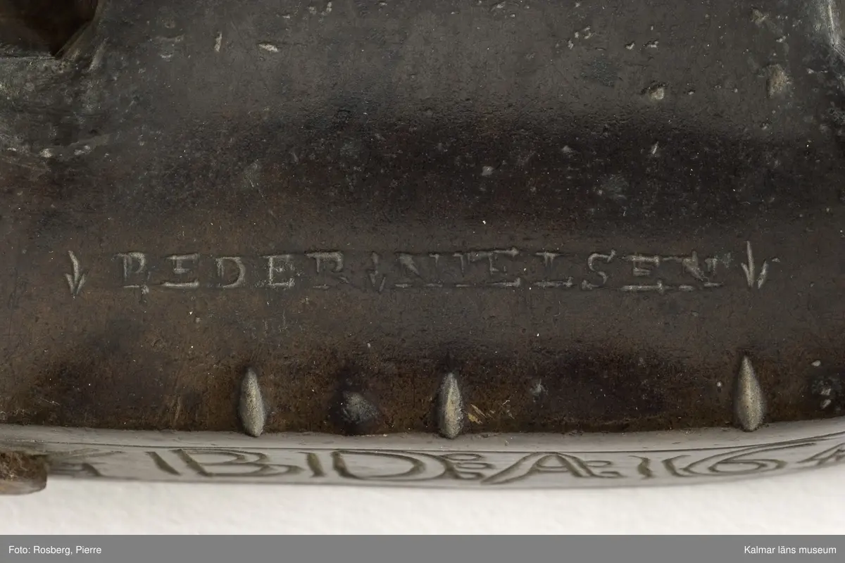 KLM 5926. Stykjärn. Av malm. Avsett för lod. Lucka baktill. Framtill rundat. Handtaget av malm. Text på smalsidan inom en ram: G.B.D.A:1647, på uppsidan inpunsat: PEDER NIELSEN. Luckan ornerad med halvmånformiga ornament,