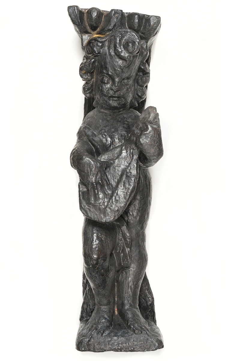 Skulptur föreställande en barnfigur med en cister (ett stränginstrument som tillhör familjen lutor).

Instrumentet hålls med bägge händer framför kroppen. Huvudet något vridet åt vänster. Kroppstyngden vilar på vänster ben medan höger ben är böjt. Utmed höger sida skymtar en mantel. Figuren står på ett minimalt underlag som troligen avser att föreställa ett stycke naturlig mark. Skulpturens baksida är slät.

Skulpturen är välbevarad.



Text in English: This is a sculpture of a child playing a cister.

It holds the instrument in both hands in front of its body. The head is turned slightly to the left. The weight of the body rests on the left leg, the right leg is bent. A mantle or cloak is to be seen on the figures right side. The child stands upon a small flat plinth. The back of the sculpture is smooth.

The sculpture is well preserved.