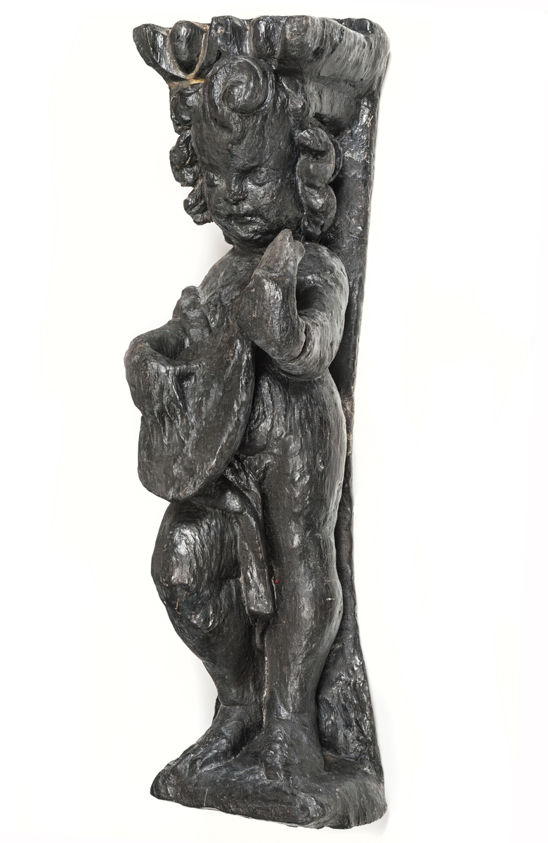 Skulptur föreställande en barnfigur med en cister (ett stränginstrument som tillhör familjen lutor).

Instrumentet hålls med bägge händer framför kroppen. Huvudet något vridet åt vänster. Kroppstyngden vilar på vänster ben medan höger ben är böjt. Utmed höger sida skymtar en mantel. Figuren står på ett minimalt underlag som troligen avser att föreställa ett stycke naturlig mark. Skulpturens baksida är slät.

Skulpturen är välbevarad.



Text in English: This is a sculpture of a child playing a cister.

It holds the instrument in both hands in front of its body. The head is turned slightly to the left. The weight of the body rests on the left leg, the right leg is bent. A mantle or cloak is to be seen on the figures right side. The child stands upon a small flat plinth. The back of the sculpture is smooth.

The sculpture is well preserved.