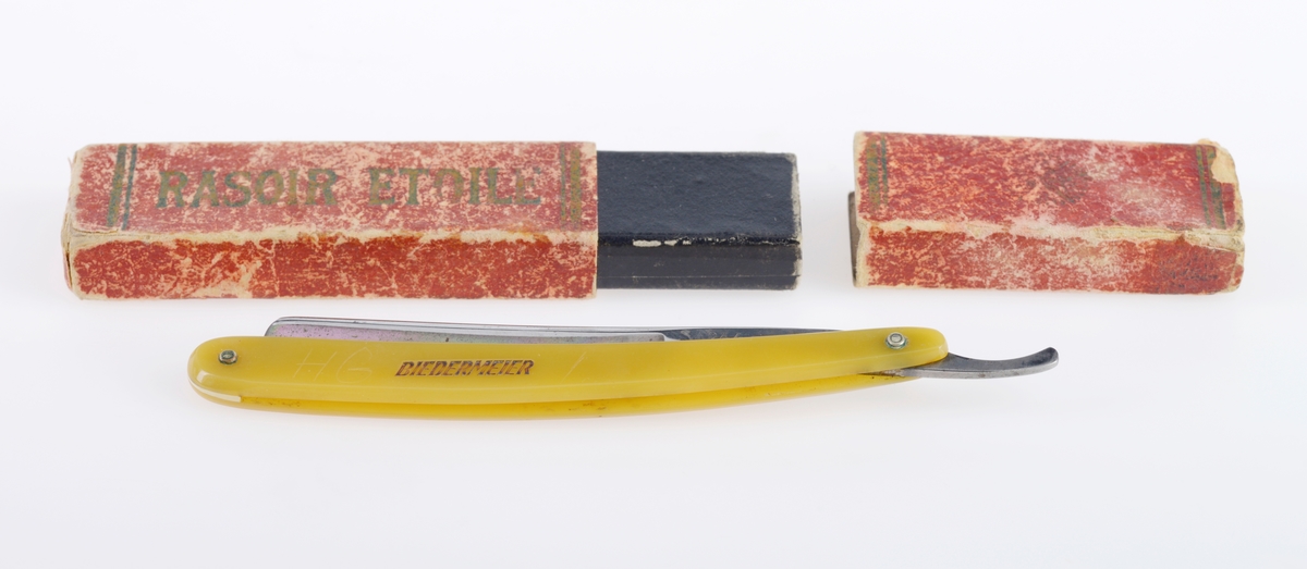 En barberkniv der kniven er laget av stål, mens håndtaket muligens er laget av bakelitt. Det er et nedfellbart skarpt blad inni et enkelt håndtak. Håndtaket er gult. Det er tekst på både knivbladet og på håndtaket, se "Påført tekst/merker". Esken er laget av papp. Utsiden er trukket med rødt papir med tekst og striper i gull. Esken er ganske slitt. Øverste del av esken kan tas av.