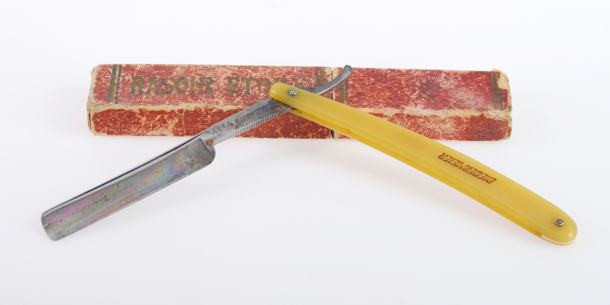 En barberkniv der kniven er laget av stål, mens håndtaket muligens er laget av bakelitt. Det er et nedfellbart skarpt blad inni et enkelt håndtak. Håndtaket er gult. Det er tekst på både knivbladet og på håndtaket, se "Påført tekst/merker". Esken er laget av papp. Utsiden er trukket med rødt papir med tekst og striper i gull. Esken er ganske slitt. Øverste del av esken kan tas av.