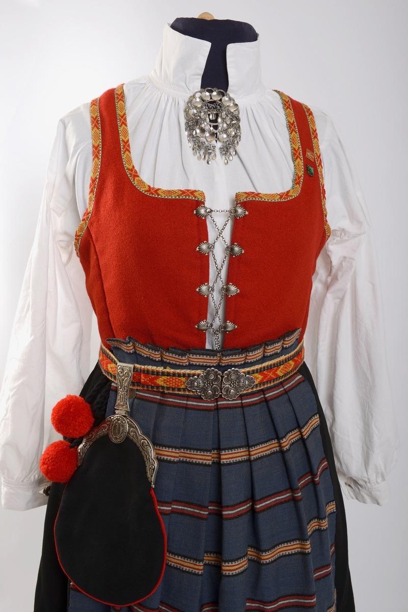 Kvinnebunad fra Vestfold, 1956 - modell.

Giver og tidligere eier, Wenche Blomberg, har fortalt dette om bunaden: 
"Vestfoldbunaden omtales i Norsk bunadleksikon b. 2, min er 1956-modellen avbildet på s.99.Jeg kjøpte den brukt på 1970-tallet en gang. Selgeren hadde sydd den selv og initialene hennes står på et lite metallmerke som er festet på innsida av capen. Husker ikke navnet. Hun bodde på Nøtterøy-sida av Kanalbrua i Tønsberg. Den passet akkurat, så jeg slapp å bruke penger på tilpassing. Jeg bare sydde på et par ekstra smålommer på den grønne understakken etter en tid. Bortsett fra snøringslenka som fulgte med, kjøpte jeg det øvrige sølvet sjøl. Lue og sko likeså. Etter noen år med heftig dansing der lua hele tida føyk av fordi jeg ikke likte å bruke hakeknyting, sydde jeg på en borrelås-strimmel inni, og da satt den bedre fast i håret. Ekstra bomullsskjorte kjøpte jeg seinere på Husfliden i Tønsberg.
 
Brukte den lite de første åra, men da jeg kom tilbake til Oslo i 1985 etter fem år i Nord-Gudbrandsdalen og etterhvert ble leikarring-aktiv, økte bruken betraktelig. I skjørtet er det ei lita rift som jeg rev opp da jeg klatra over et nettinggjerde i hagen utafor bygningen som tidligere huset Norges geografiske oppmåling, seinere Kunstakademiet, der det ofte var forskjellige festlige arrangementer. Jeg husker ikke anledningen og hvorfor jeg ble hengende fast i det gjerdet, men skaden var ikke større enn at jeg reparerte den sjøl. Pompongene på forklesnora var ellers et sårt punkt, måtte få noen til å lage nye for meg en gang, men jeg husker ikke hvem som gjorde det - kanskje Husfliden ett eller annet sted. Låsen på lauslomma var også til stadig irritasjon fordi den sprang opp hele tida så tobakken min datt ut.

Når man er folkedanser er skoa nærmest hjertet! Disse skoa var til slutt så godt inntrødde at jeg kvidde meg for å bruke et par nye jeg kjøpte via BUL. (Forresten: Hvilket par fikk dere? De gamle slitte, eller de nye? Begge?) [Opplysning fra Kvinnemuseet: Bare de nye skoene er del av gaven]

Seinere på 80-90-tallet en gang fikk jeg lagt stakken ut i livet av systova på Heimen. De bildene jeg har funnet er fra tida i Leikarringen i BUL. Det er forresten ikke lett å ri med bunadsstakk, kan jeg meddele. Hvor skal man gjøre av alt stakkestoffet der man sitter overskrevs? 

Jeg brukte bunaden siste gang 17. mai 2017 i Svullrya. Da hadde den krympa så mye at jeg knapt fikk den på meg, og nå så jeg ingen grunn til å bruke mer penger på utvidelser."

Til bunaden hører også to jakkemerker, ett fra Noregs ungdomslag, NU, og ett med symbol av en drage, som er merket for mikrostaten Ladonia, der Wenche Blomberg er Minister of Madness og Enkegrevinne Wenche von Blomberg und Grünerløkka. 

Om det siste merket forteller Wenche Blomberg: 
"Ladonia: Virtuell ministat opprettet av kunstneren Lars Vilks. Det finnes en god del slike på nettet, men Ladonia er i tillegg knyttet til et konkret geografisk område ved Kullen i Skåne: ei nesten utilgjengelig strandstripe av kjemperullestein der Vilks har bygd opp en rekke monumentale skulpturer av rekved. Historie, bilder etc på www.ladonia.org. Det har forekommet åratals med fornøyelige tvister med grunneiere og staten Sverige. Alle kan bli borgere av Ladonia, noe som fra tid til annen har ført til massehenvendelser fra en del land i Asia fra folk som vil emigrere og søker visum. Statsbudsjettets inntektsside stammer i stor grad fra salg av titler. Det var sånn jeg ble enkegrevinne og står i adelskalenderen. Man kan foreslå seg selv til minister for et hvilket som helst fantasirikt fagområde, men dette må godkjennes av regjeringen. Det var ingen innvendinger mot min søknad om å bli «minister of madnesss", man fant at den passet utmerket inn."
