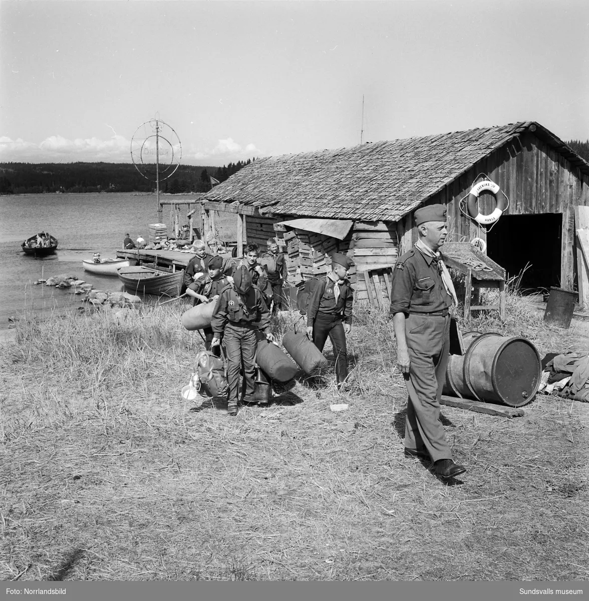 I juli 1959 anordnade Svenska Scoutförbundet sitt stora internationella läger på Rödön utanför Alnö. På första bilden ses lägerledaren Tage Allegård anlända med en grupp pojkscouter för några intensiva veckor på ön. Lägret hade förberetts under ett helt år och dagarna innan hade 70 pojkscouter och ledare hjälpts åt att bygga upp de olika lägerbyarna, som fick namn efter olika fyrar.  SCA hade upplåtit en pråm som låg för ankar i hamnen fylld med de matvaror som krävdes för att mätta de cirka 800 deltagarna.