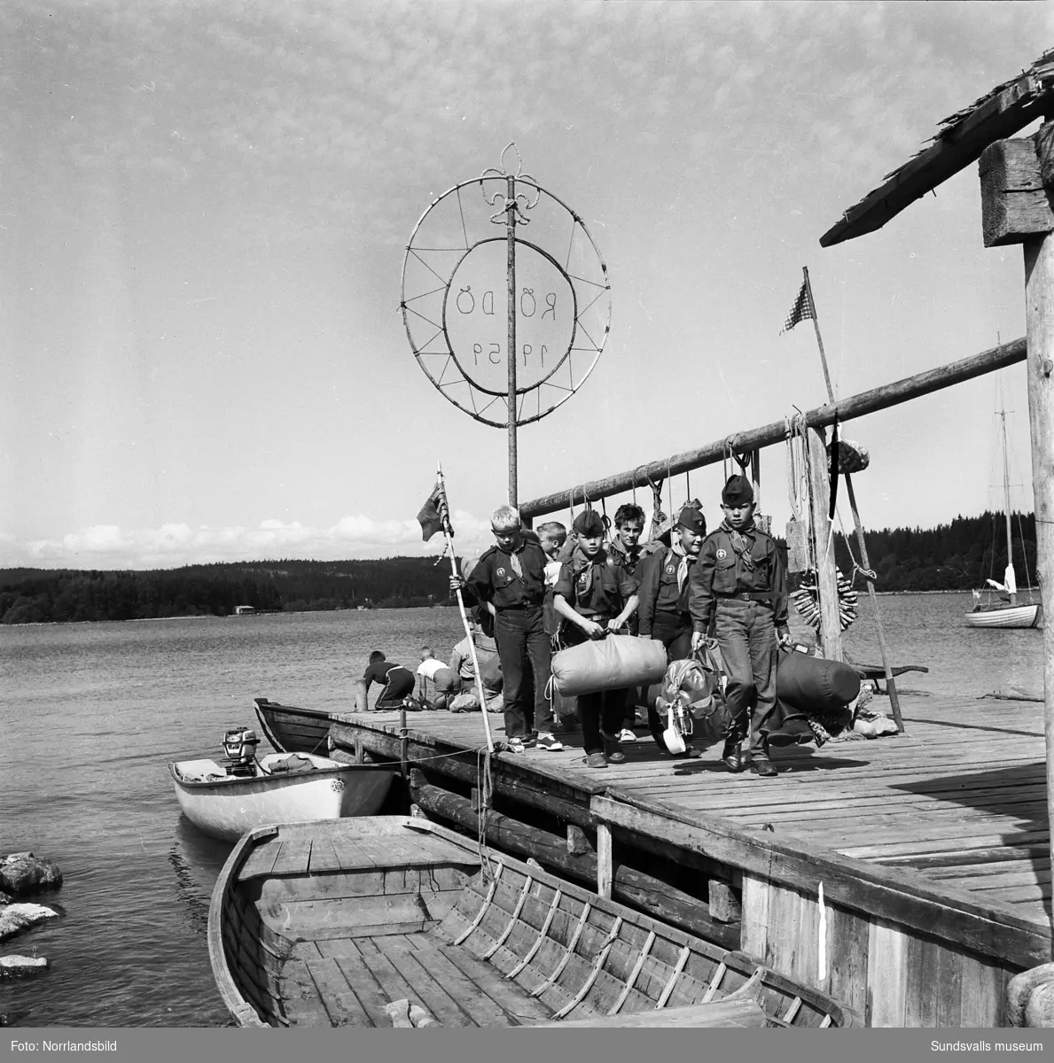 I juli 1959 anordnade Svenska Scoutförbundet sitt stora internationella läger på Rödön utanför Alnö. På första bilden ses lägerledaren Tage Allegård anlända med en grupp pojkscouter för några intensiva veckor på ön. Lägret hade förberetts under ett helt år och dagarna innan hade 70 pojkscouter och ledare hjälpts åt att bygga upp de olika lägerbyarna, som fick namn efter olika fyrar.  SCA hade upplåtit en pråm som låg för ankar i hamnen fylld med de matvaror som krävdes för att mätta de cirka 800 deltagarna.