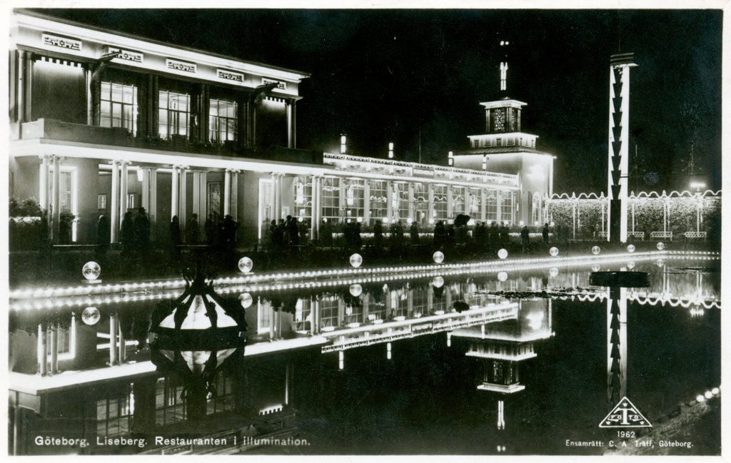 "Liseberg, Restauranten i illumination."
En byggnad med stora fönster och upplyst fasad speglar sig i en spegelblank damm. Människor står framför byggnaden med syns endast som svarta skuggor, fotografiet taget kvälls- eller nattetid.