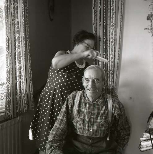Hildings fru kammar håret på en man som sitter i en stol framför henne, 1969.