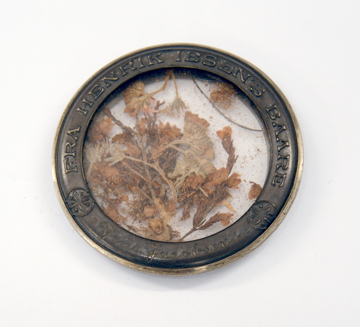 Båremedaljong fra Henrik Ibsens båre. Pressa blomster i en medaljong av sølv og glass. I hvitt etui. Sølvramme med blomsterdekor på en side og tekst på den andre siden: FRA HENRIK IBSENS BAARE. Jens Jacobsen. Innvendig i etuiet bokstavene hi og under etuiet J.Tostrup, Kristiania.