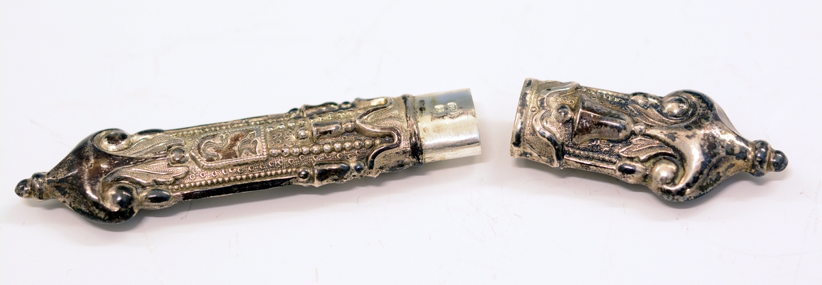 Nålehus i sølv. Drevet dekor, bl.a. med bjeller og våpenskjoldform med fugl og slange. 
Utydelig stempel: Ant. krone og (...)114.