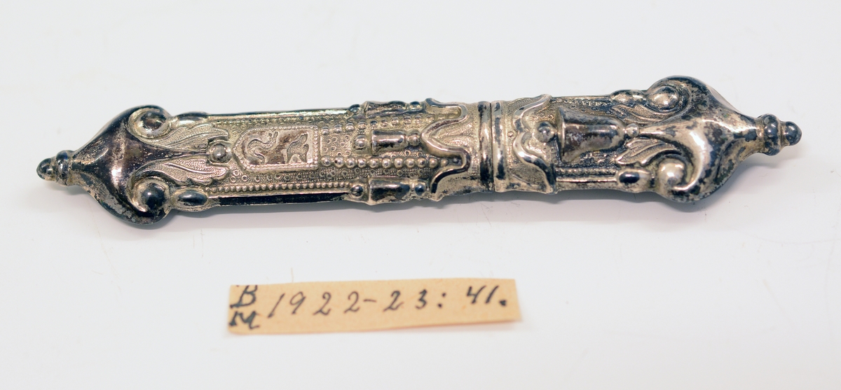 Nålehus i sølv. Drevet dekor, bl.a. med bjeller og våpenskjoldform med fugl og slange. 
Utydelig stempel: Ant. krone og (...)114.