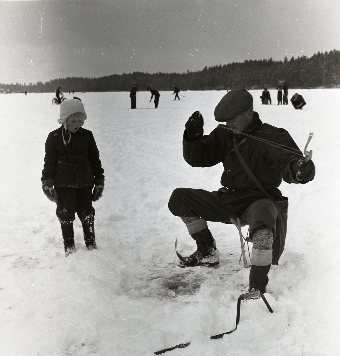 En man sitter vid ett pimpelhål och drar upp en fisk. Ett barn står bredvid och tittar på. IOGT och FK pimpeltävling på Skidtjärn 14 april 1957.