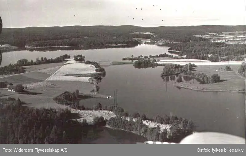 Oversiktsbilde over del av Nipelva med Nipa bru i Varteig 1951. Brua forbinder Hafslundsøy (Tune) og Varteig, går over Nipelva, en gren av Glomma. Påbegynt bygget i 1914 og ferdig i 1917. Brua ble sprengt i april 1940. Bygget opp og åpnet i 1941. Hasle gård, gn.nr.1, b.nr.1, i Varteig til venstre.