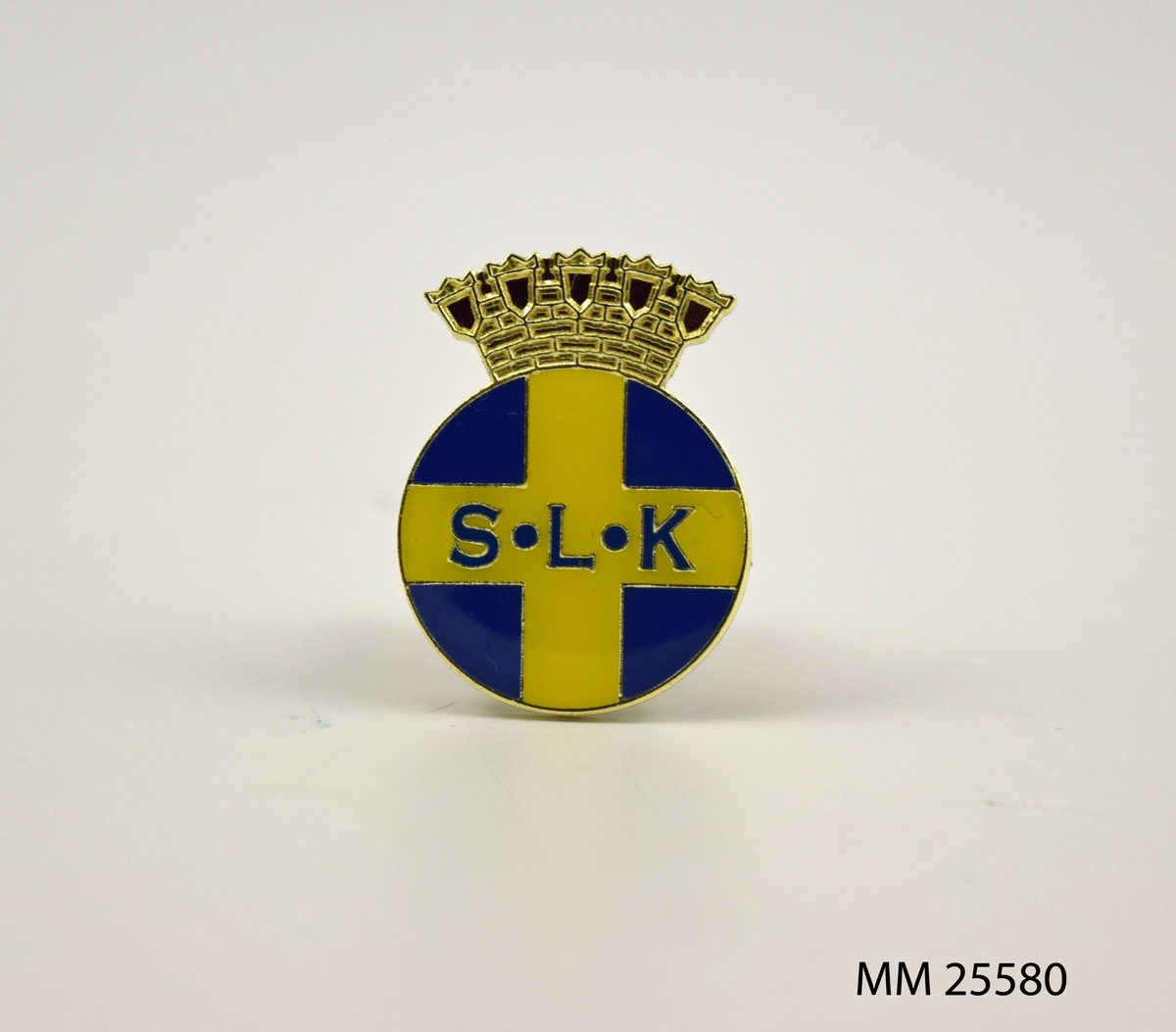 Lottamärke av metall. Cirkelrunt märke med krona i guld över. Emaljerat motiv av svenska flaggan, blå bakgrund med gult kors. Bokstäverna "SLK" i blått i korsets mitt. Baktill rund magnet för att fästa märket på kläder.