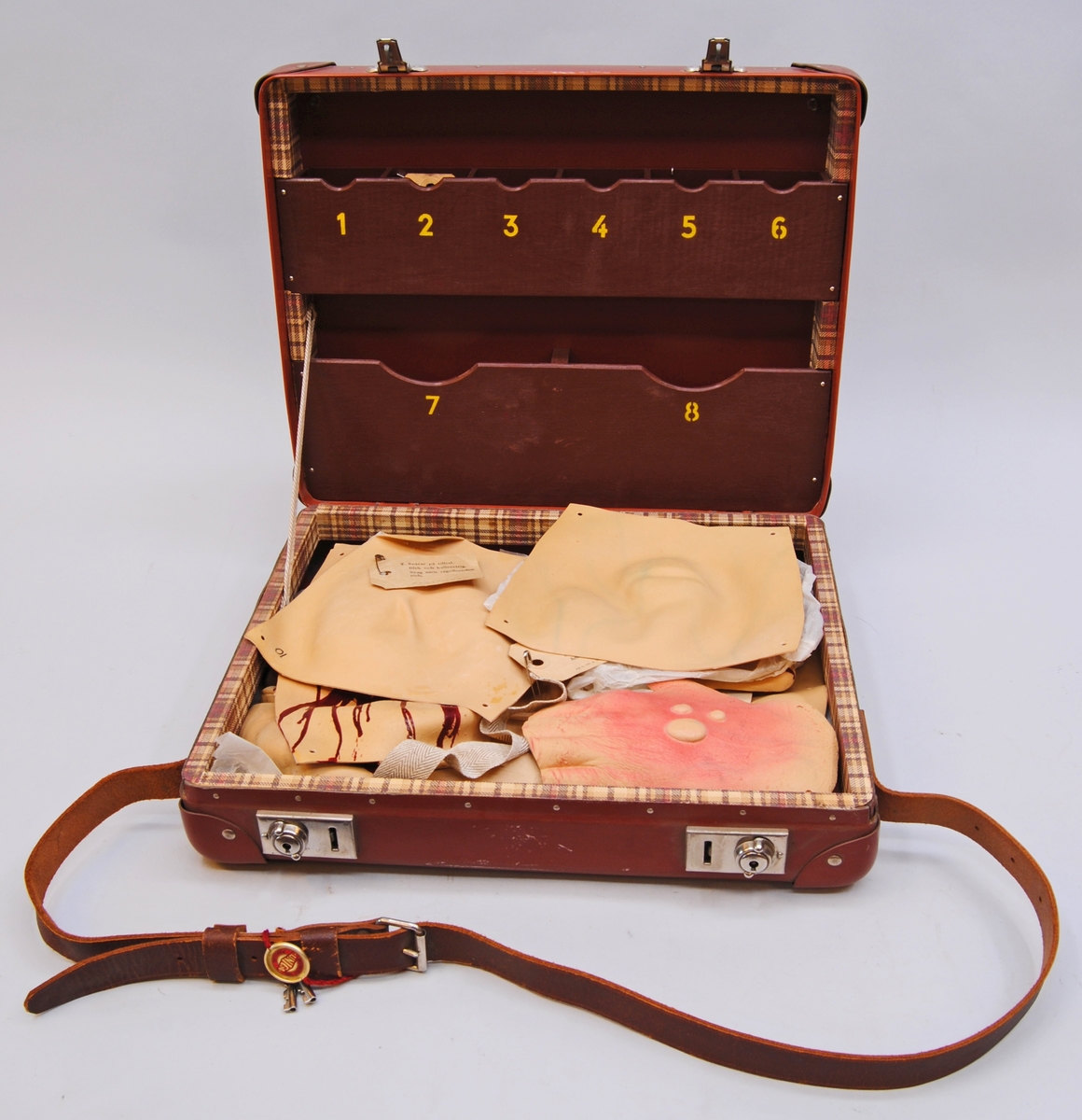En utbildningsväska av handkofferttyp, gjord av hårdpapp och lackad i roströd färg. På väskans sidor sitter läderförstärkningar i form av infärgat läder fastsatta med metallnitar. Väskan har axelband av brunt läder samt två låsbara lås av metall. Två nycklar i rött band hänger på axelbandet, på bandet återfinns en "metallsigill" med märkningen "Aktiebolaget Tidan Unica Mariestad".
Väskan är inredd med en träram klädd i rutigt papper. Den övre inredningen består av två fackdelar av trä, även det i roströd färg. De övre facken består av sex olika fack, den nedre av två större. Facken är märkta med siffror i gult tryck.
Väskan har även två spännband för att innehållet ska hållas på plats.

Väskan innehåller 25 stycken sårattrapper av gummi som imiterar diverse skador. 
1 för ansikte (:1)
1 för öra (:2)
1 för hand (:3)
2 för fot (:4-5)
20 stycken för oidentifierbara delar av kroppen. (:6-25)
1 kuvert med innehåll (:26)
1 väska (:27) 
6 lappar med beskrivning av de olika skadorna finns för att fästa på attrapperna. Varje attrapp har en märkning i form av nummer som sätts ihop med lapparna. En av attrapperna har tillverkarnamn tryckt på baksidan. Det finns också gummiband, resårband och metallhyskor för fästning av attrapperna i lådan, liggandes i ett kuvert märkt med tillverkarnamn. Handledningsmanual från Röda Korset återfinns det med.