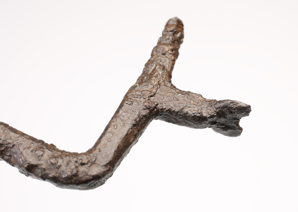 Rund böjd ten som avslutas med en ändknopp i ena änden. Anda änden rätvinkligt böjd och överst gängad och försedd med ett vinkelrätt utskott. Detta utskott är avslutat med en nu ofullständig rund platta med hål. Troligen en avtryckare till ett armborst eller annat handvapen.