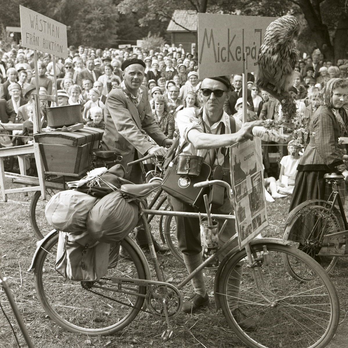 Ett ekipage som deltar under karneval vid hembygdsfest i Rengsjö. Ekipaget består av två män på cykel med packning på pakethållaren, varsin banderoll med text och bild på, 22 juni 1962.