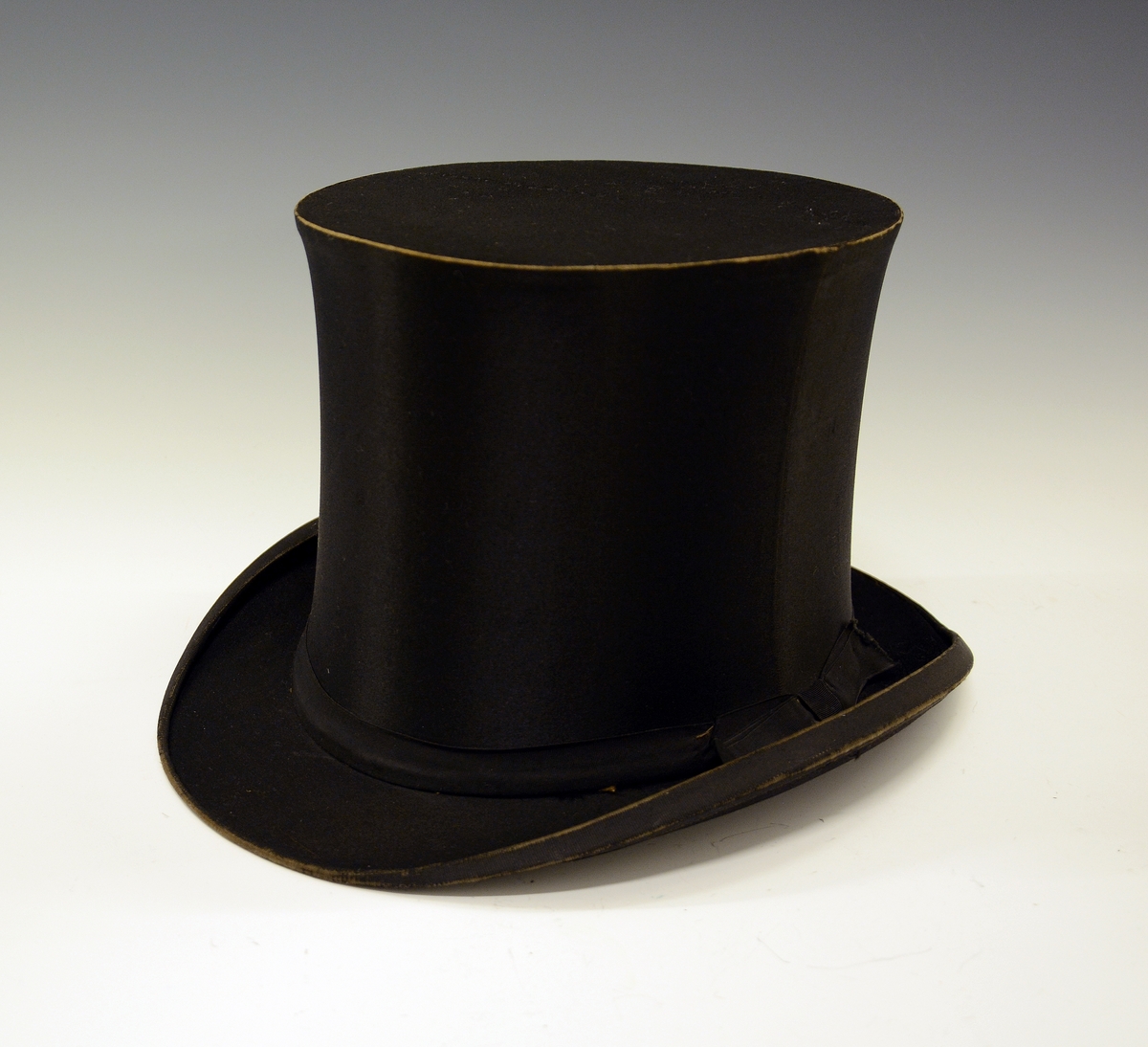 Flosshatt med etui. Sort, sammenleggbar flosshatt eller Chapeaux claque. Silketrukket papp med bånd nederst. Monogram HI med metallbokstaver innvendig. Ovalt etui i brun papp med påskrift i gull: "MANUFACTURE DE PARIS CHAPEAUX MÉCANIQUES PERFECTIONÉS", samt våpenskjold med blant annet skip i midten. Strikk og metallfester på sidene. Kjøpt av firmaet L.H.van Hees, München. Fabrikkert av Giraud, Paris.