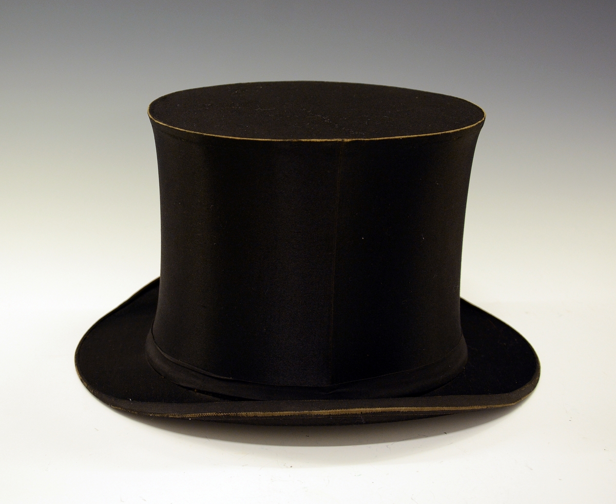 Flosshatt med etui. Sort, sammenleggbar flosshatt eller Chapeaux claque. Silketrukket papp med bånd nederst. Monogram HI med metallbokstaver innvendig. Ovalt etui i brun papp med påskrift i gull: "MANUFACTURE DE PARIS CHAPEAUX MÉCANIQUES PERFECTIONÉS", samt våpenskjold med blant annet skip i midten. Strikk og metallfester på sidene. Kjøpt av firmaet L.H.van Hees, München. Fabrikkert av Giraud, Paris.
