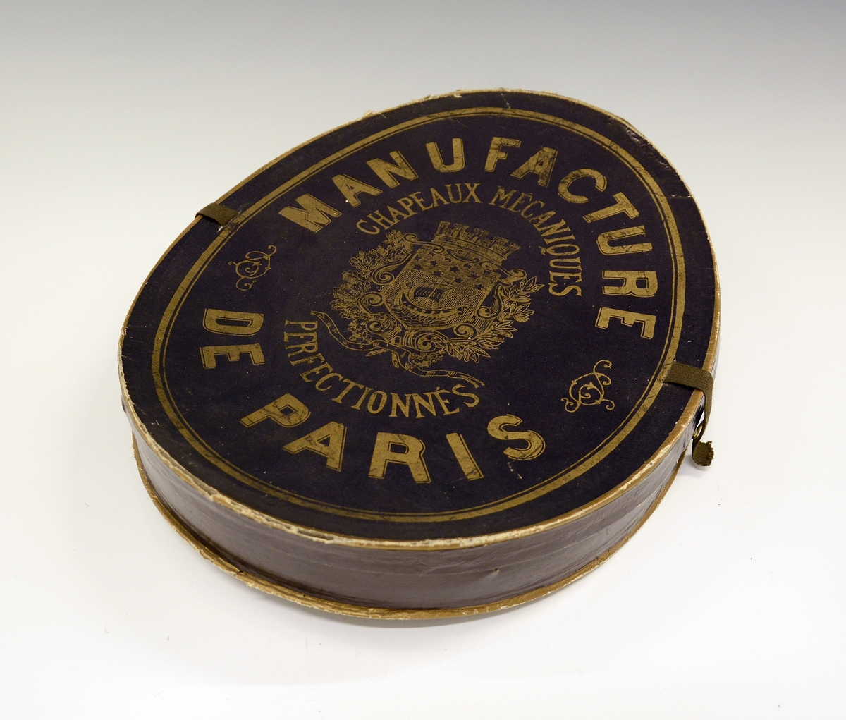 Flosshatt med etui. Sort, sammenleggbar flosshatt eller Chapeaux claque. Silketrukket papp med bånd nederst. Monogram HI med metallbokstaver innvendig. Ovalt etui i brun papp med påskrift i gull: "MANUFACTURE DE PARIS CHAPEAUX MÉCANIQUES PERFECTIONÉS", samt våpenskjold med blant annet skip i midten. Strikk og metallfester på sidene. Kjøpt av firmaet L.H.van Hees, München. Fabrikkert av Giraud, Paris.