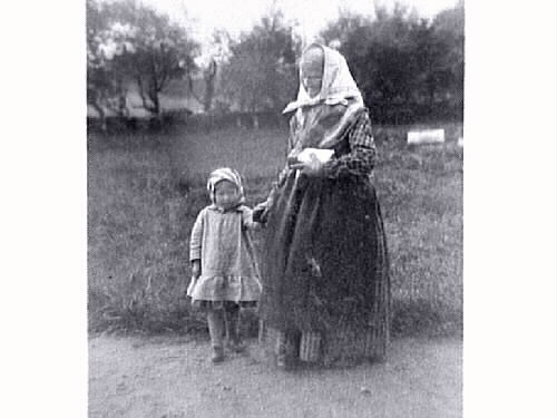 "Med mormor till kyrkan år 1890"
E5804_1: Gammal kvinna står utomhus med ett litet barn vid ena handen och en tjock bok i den andra, troligen en bibel då de på bild nr 2 sitter i Tjärby kyrka.
E5804_2: Gammal kvinna och litet barn sitter på en utsirad bänk i Tjärby kyrkas kor. Man ser lite av altartavlan och altarringen.