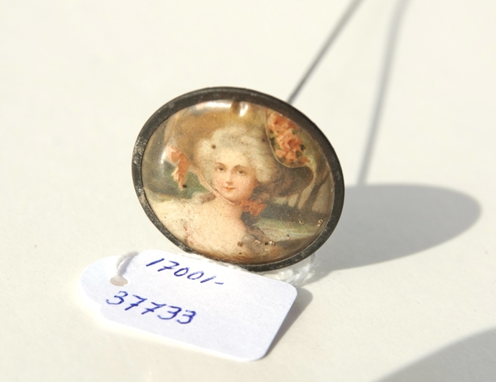 Hattnål i metall. Rund knapp med bild på dam med vitt hår, stor hatt med blommor, fäst mot knappen med smal kant av metall. Guldfärgad baksida med metallsläpp. Lång smal nål för fastsättning i hatten.