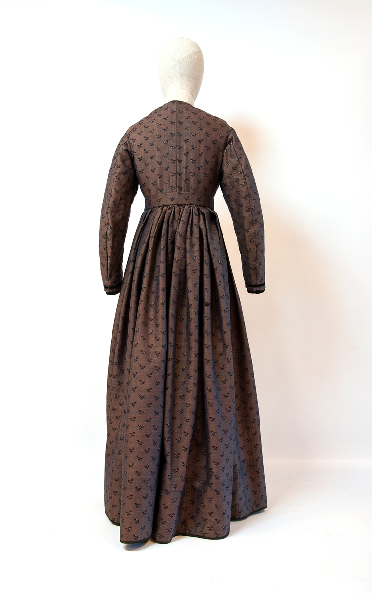 Kjole sammensydd av liv og skjørt med belte i livet med sløyfe i samme stoff. Halvull verkenstoff med trykte blomster. Sydd i Sørum i Akershus ca 1860 og senere brukt på Hamar. Skjørtet har mye håndsøm men vi ser også maskinsøm. Skjørtet er fóret med en skoning av lin i nedre kant. Det er også festet et grønt smalt bånd som slitekant. Overdelen er fóret med ufarget linstoff. Svart fløyelsbånd er festet 1,5 cm fra ermekanten.