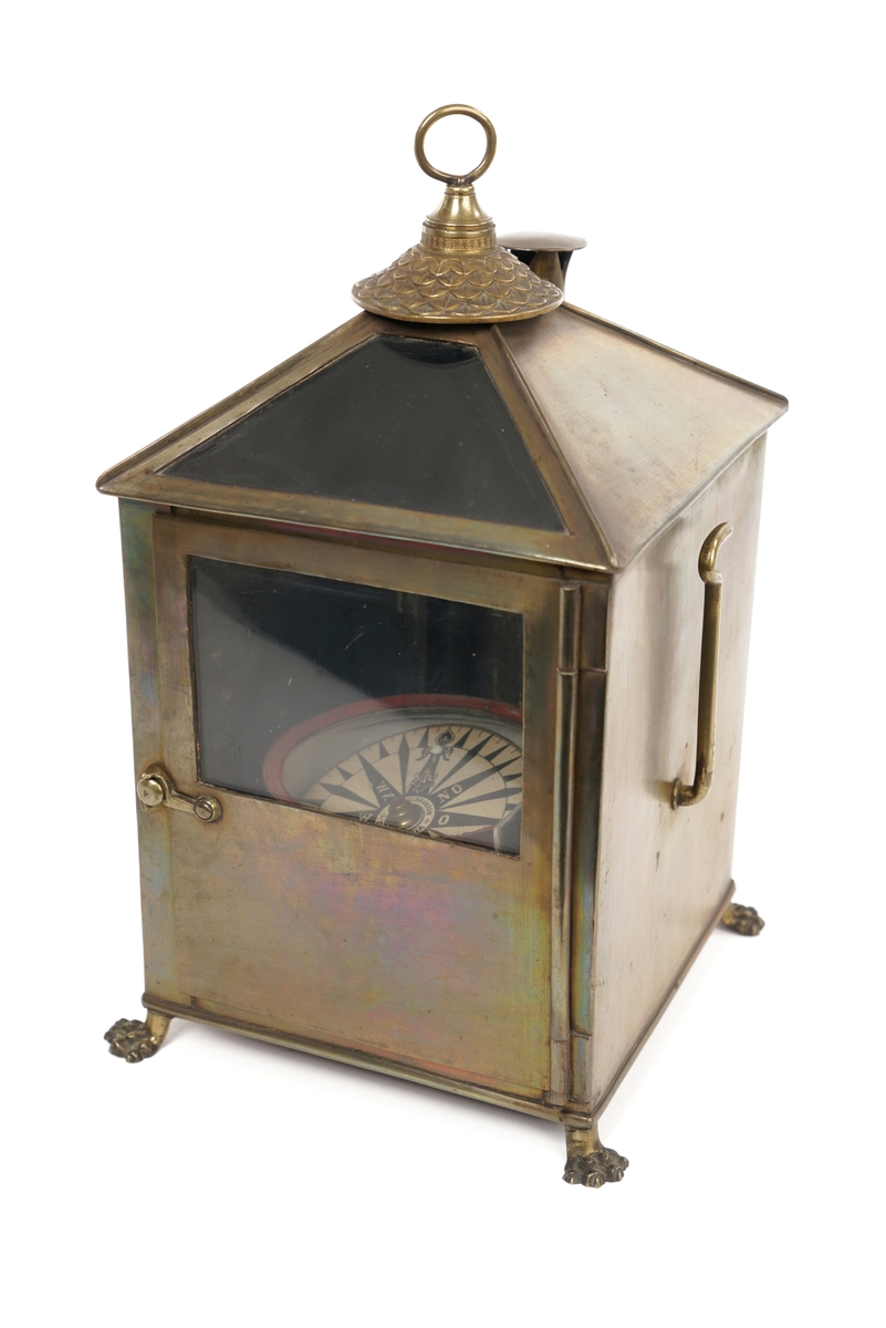 Firkantet kompasshus av metall med dør med vindu av glass. Døren kan låses med krok. Fire forsirede føtter av metall.  Tak som skråner til alle fire sider med vindu på takets forside. Luftavtrekk. Toppen av taket er utstyrt med forsiret deksel med bærering. To støttekroker, èn på hver side. Inne i huset er det to stearinlysholdere med metallfjær-innretning. Mellom disse er det fastmontert et metallrør.
Videre medfølger det to støtteklosser av tre for kompasset. Kompasslokk følger også med.