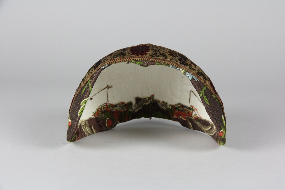 Bindmössa av hårt stärkt textilstomme. Klädd med blommig brokad med brun botten. Runt mössan är ett brett blommigt sidenband fäst med nålar.