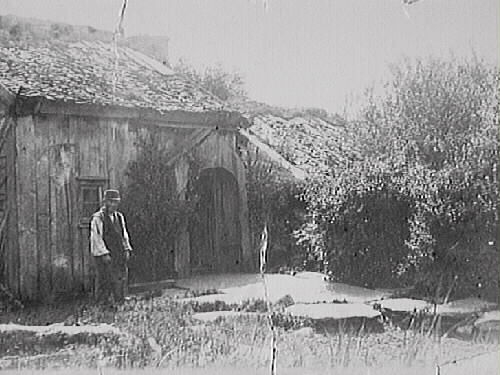 Ryggåsstuga med bislag för ingången, s k skunke (skånke). Framför huset är marken belagd med stenhallar och där står en man i väst och keps. Bilden märkt "Carl Heintz".