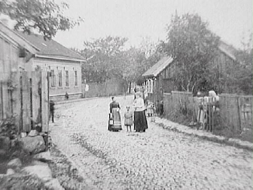Stadsgata med kullersten utmed ett långt trähus med trädgård på vänster sida och ett staket på höger sida. I mitten står två kvinnor och ett barn på gatan och vid grinden står ytterligare en kvinna.