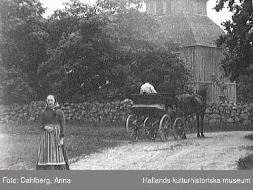 Okome gamla träkyrka. I förgrunden står en kvinna och bakom henne rullar ett hästekipage bortåt på den sandiga vägen.

 Se Information, även tidigare bildnr.