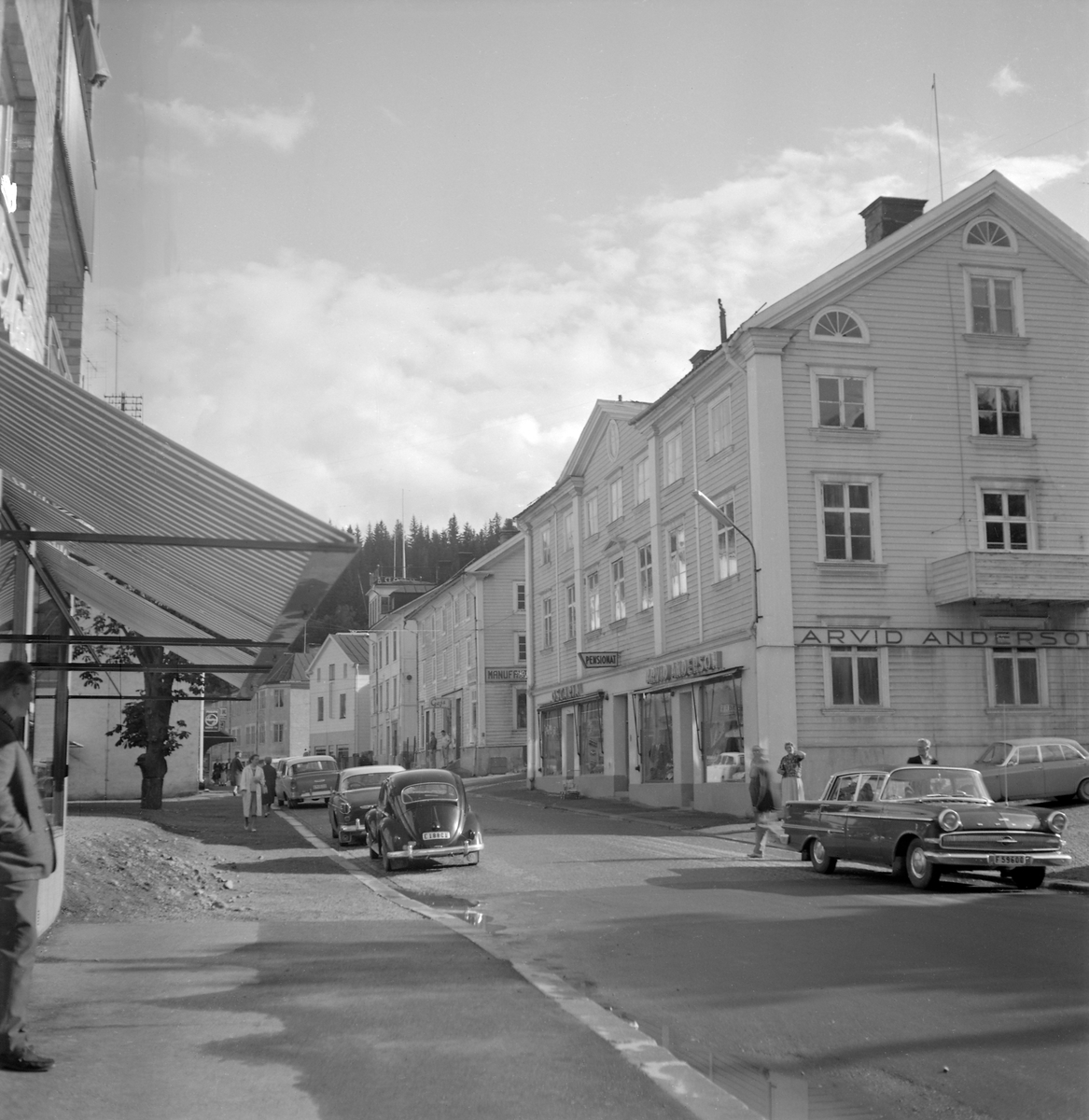 Parti av Storgatan i Valdemarsvik 1962. Inom kort kommer trähusen på gatans högra sida att rivas och ersättas med tidstypiska affär- och bostadskomplex. Närmast ses vad samtiden kallade Centrumhuset, inrymmande sina sista hyresgäster i skobutiken Oscaria, Arvid Anderssons Herrekipering och pensionat Centrum. Byggnaden var uppförd 1849. I bortre änden av den rivningsbeslutade husraden skymtar Stora hotellet.