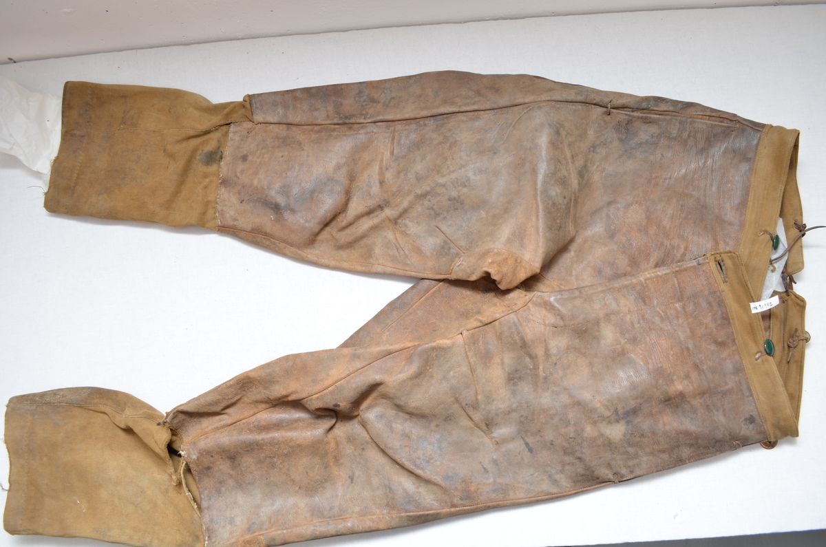 Brun skinnbrok (langbrok). Nedre del av buksebeina er i brunt bomulltøy. (26 cm). (Kan det ha vore kortbrok tidlegare? ) Linning og for i gylfen er i same bomullstøyet.To stikklommer framme  med blått bomullstøy på innsida. Gylf. Open saum midt bak (7 cm), hekta saman med ei skinnreim. Forskjellige knappar.