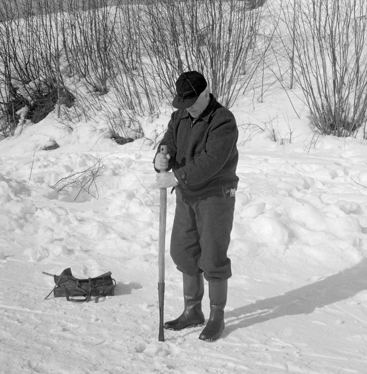 Leiv Ørbak fra Elverum hakker hull i elveisen på Glomma nedenfor Gammelbrua i Elverum, der han drev med «grindalsfluefiske».  I dette spesielle isfisket nyttiggjorde man seg en spesiell steinflueart (Capnia sp.) som lokkemat.  På nyfestadiget lever «grindalsdflua» eller «sprela», som den også kalles, i vann.  Når sollyset blir kraftigere utover våren søker nymfene mot lyset i sprekker i isen, der de etter hvert kravler seg opp i dagen, hvor de utvikler vinger og blir til små fluer.  Det er som nymfer at dette insektet er interessant for fiskerne.  De hogger derfor hull på isen, som de fyller med et tynt snølag, for å hindre at fluene kommer seg helt opp.  Snøen med alle nymfene kan de etter hvert samle i et spann.  Under fisket slipper de nymfer ned i fiskehullet eller et ovenforliggende hull, der de tiltrekker seg beitende sik og harr.  Fiskerne la seg deretter på magen, med et eller annet underlag på isen, og blikket vendt ned i vannmassene.  Der fisket de, vanligvis med et snøre med en krok med meitemark som agn, men i noen tilfeller også med en tynn messingtråd med ei renneløkke i enden.