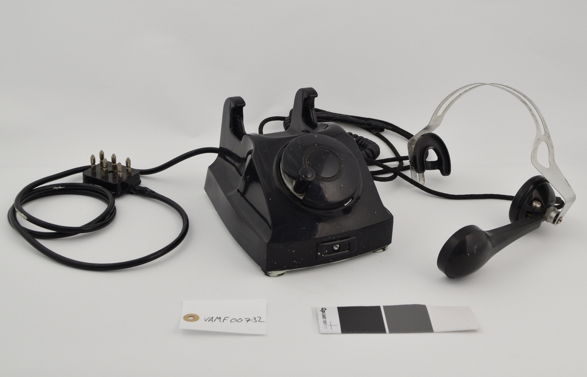 Telefonapparat med telefonrør festet til en hodebøyle i metall med ledning i mellom rør og apparat. Apparatets grunnflate er avlang heksagonformet (sekskantet). Skråner nedover foran der det er plassert en rund nummerskive som er dekket av et rundt deksel med sveiv. Skråner ned bak der gaffelen er plassert, med plass til å plassere hodebøylen med røret. En hodemikrotelefon er koblet til apparatet og har erstattet det originale telefonrøret. Hodemikrotelefonen består av en hodebøyle med høyttaler i venstre øre, og det er der festet en mikrofon fra denne i et ledd som gjør den justerbar.