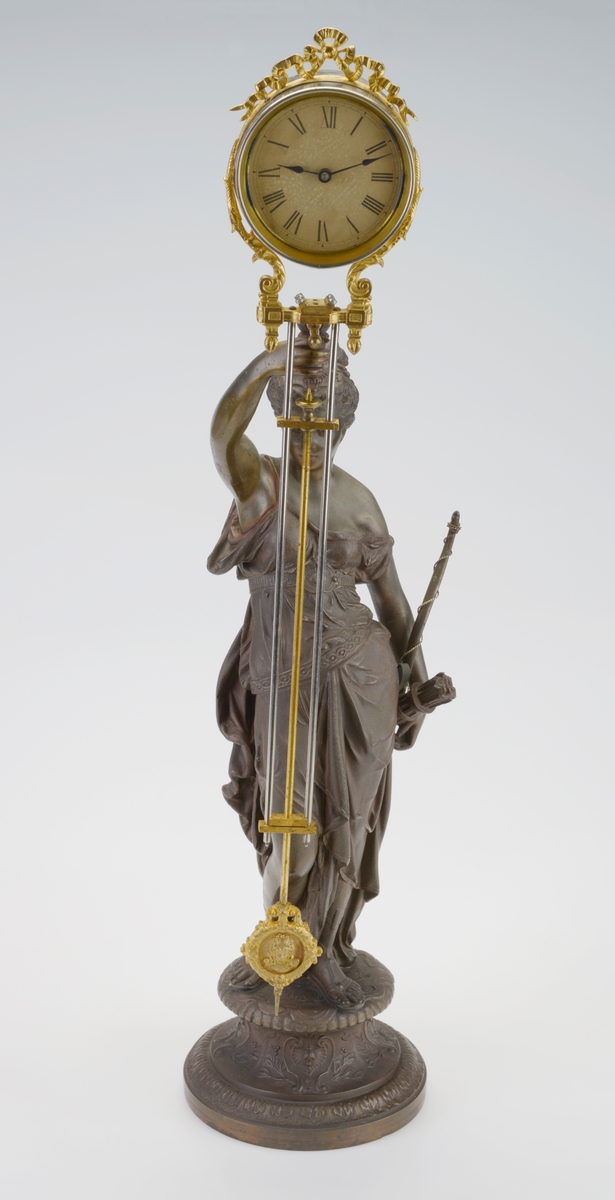 En pendelklokke til å ha stående på et bord, en hylle eller lignende. Den består av en figur/liten statue og en pendel med urverk. Figuren er av støpt metall som er påført maling/lakk slik at det ligner på bronse. Figuren står på en 9,5cm høy sokkel. Det er støpt dekor inn i sokkelen. Figuren forestiller Diana som i romersk mytologi var jaktens gudinne og assosiert med ville dyr, skogen og månen. Hun har en løstsittende kjole med belte og på beina har hun sandaler. Håret er satt opp og i front har hun en kam/liten tiara. Ved den ene hånden har hun bue med sølvfarget metalltråd tvunnet rundt. På denne siden henger det også en kogge med piler. Den andre hånden holder hun over hodet og holder en gjenstand som pendelen skal henges på. Pendelen er laget av forgylt messing og metall. Øverst er det en rund urkasse av blankt metall som er festet i forgylt "messinggrep". Urskiven viser tall fra romertall I - XII. Viserne er laget av svartlakker metall. På urskiven er det informasjon om produsent, se "Påført tekst/merker". På baksiden kan man se urverket som er laget av messing (visst nok et 8-dagers urverk). Fra festet til urkassen henger det to sølvfargede metallstenger med en messingfarget metallstang i midten. Nederst på pendelen er det en utsmykket ramme med et et kvinneportrett i. Dette er støpt og gullforgylt.