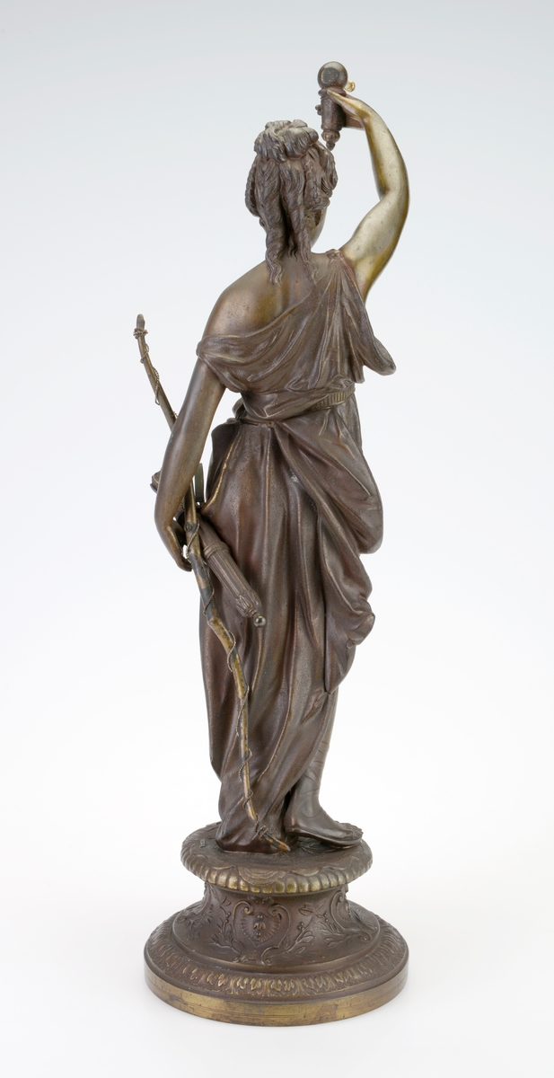 En pendelklokke til å ha stående på et bord, en hylle eller lignende. Den består av en figur/liten statue og en pendel med urverk. Figuren er av støpt metall som er påført maling/lakk slik at det ligner på bronse. Figuren står på en 9,5cm høy sokkel. Det er støpt dekor inn i sokkelen. Figuren forestiller Diana som i romersk mytologi var jaktens gudinne og assosiert med ville dyr, skogen og månen. Hun har en løstsittende kjole med belte og på beina har hun sandaler. Håret er satt opp og i front har hun en kam/liten tiara. Ved den ene hånden har hun bue med sølvfarget metalltråd tvunnet rundt. På denne siden henger det også en kogge med piler. Den andre hånden holder hun over hodet og holder en gjenstand som pendelen skal henges på. Pendelen er laget av forgylt messing og metall. Øverst er det en rund urkasse av blankt metall som er festet i forgylt "messinggrep". Urskiven viser tall fra romertall I - XII. Viserne er laget av svartlakker metall. På urskiven er det informasjon om produsent, se "Påført tekst/merker". På baksiden kan man se urverket som er laget av messing (visst nok et 8-dagers urverk). Fra festet til urkassen henger det to sølvfargede metallstenger med en messingfarget metallstang i midten. Nederst på pendelen er det en utsmykket ramme med et et kvinneportrett i. Dette er støpt og gullforgylt.