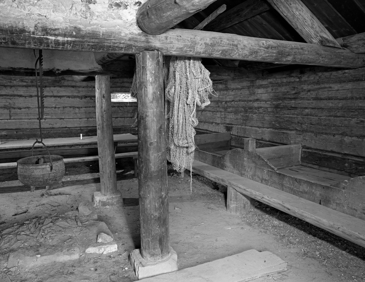 Interiør fra den gamle «Knuvelbua», som opprinnelig stod ved fiskevarpet Knuvelen i Gudbrandsdalslågen, en grusbanke nokså rett vest garden Storhove i Fåberg.  Bygningen er seinere demontert og flyttet til friluftsmuseet på Maihaugen i Lillehammer by, der den er plassert ved «Breisjøen».  Har framstår Knuvelbua som en laftet bygning med jordgolv.  Interiøret er enkelt.  Det besto av faste senger med en halvkløvningsbenk langs den ene veggen og et langbord langs den andre.  Midt i rommet var det et åpent ildsted på en lav elvpall av tilhogd stein.   Omkring dette sto det fire stolper (hvorav to er synlige på dette fotografiet), som støtter for krysningspunktene mellom tverrgående beiter og langsgående slinder i rommets raftsone.  Oppå disse var det murt et pipeløp.  Da dette fotografiet ble tatt hang det ei skjerding med ei støpt jerngryte over ildstedet og det hang et bundet fiskeredskap (ei not) på ei stang ved siden av ildstedet.  På veggen i bakgrunnen skimtes en smal lysåpning.  Bygningen ble opprinnelig brukt som kvilested for menn som deltok i det store sesongfisket etter lågåsild (Coregonus albula) i den nedre delen av Lågen.