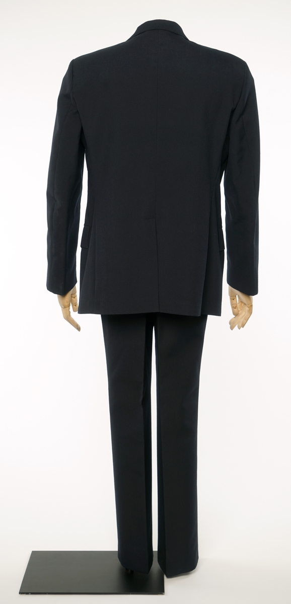 Mørkblå jakke enkelspent med fire riksløveknapper. Liggende krave med en eiksblade distinksjon med gullbånd på hver side. Stoffet er et enkelveved stoff, antatt en blanding av ull og syntetisk fiber. Jakke er foda med antatt syntetisk stoff, mørkblå. 
Bukser av samme stoff.