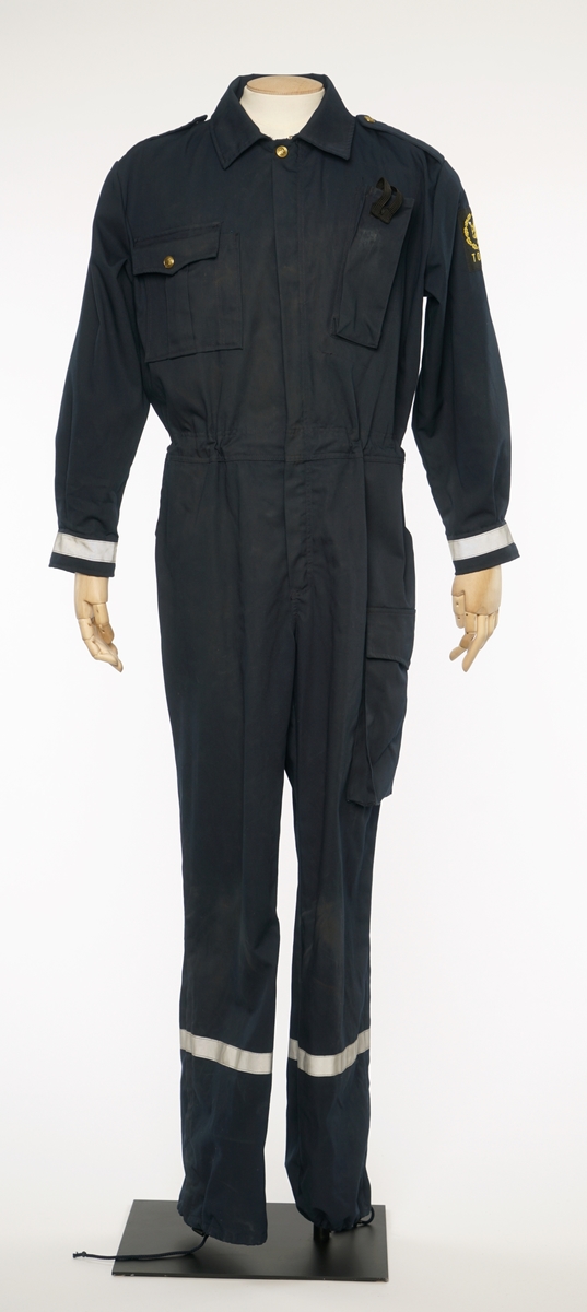 Mørkblå kjeledress med sølv refleksbånd på bukser og mansjetter. Epauletter uten tilsatt distinksjoner. 'TOLL' is refleks bokstaver på rygg. Tekstil merke på venstre arm, Riksløve med hvit øks omgitt av eikeblader over TOLL. Hempe i nakke. Kypert bomull stoff.
Nøkkelring med lær anheng funnet i høyre lomme.