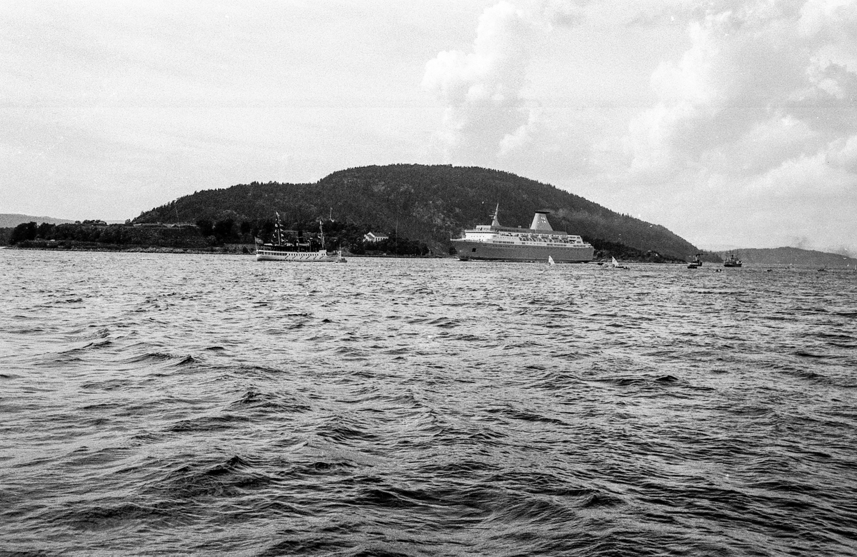 Drøbaksfjorden, sundet ved Oscarsborg. Tre menn på brygga ser på båtene med kikkert. Sandtaket og småhus på Hurum.
Fotograf: ØB Rønning
