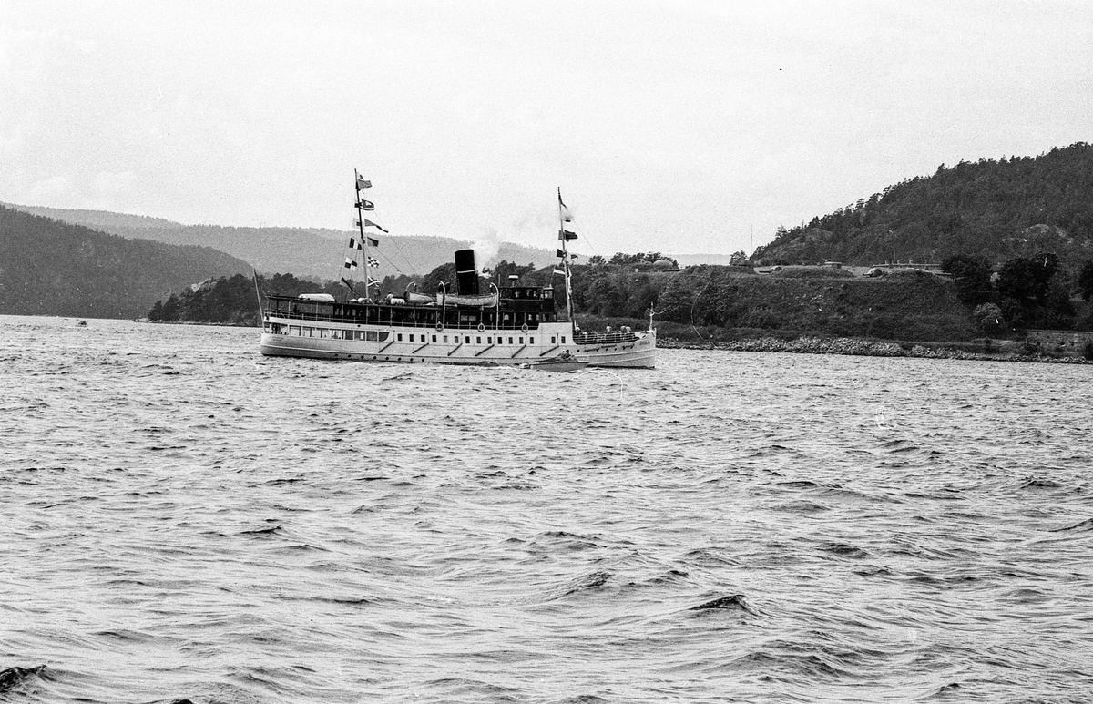 Drøbaksfjorden, sundet ved Oscarsborg. Tre menn på brygga ser på båtene med kikkert. Sandtaket og småhus på Hurum.
Fotograf: ØB Rønning