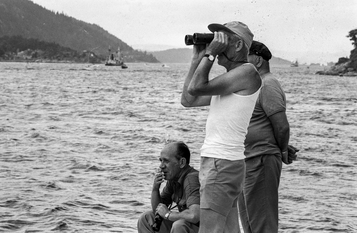 Drøbaksfjorden, sundet ved Oscarsborg. Tre menn på brygga ser på båtene med kikkert. Sandtaket og småhus på Hurum.
Fotograf: ØB Rønning