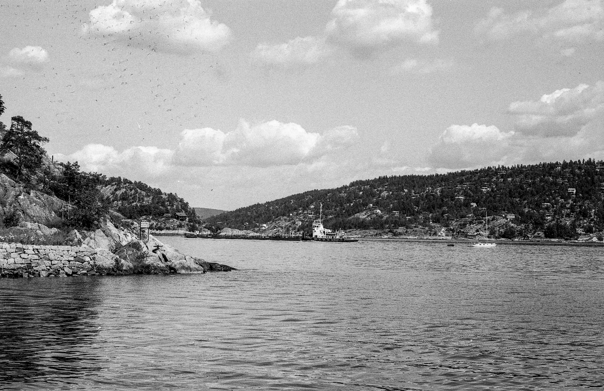 Drøbaksfjorden, sundet ved Oscarsborg. Tre menn på brygga ser på båtene med kikkert. Sandtaket og småhus på Hurum.
Fotograf: ØB Rønning