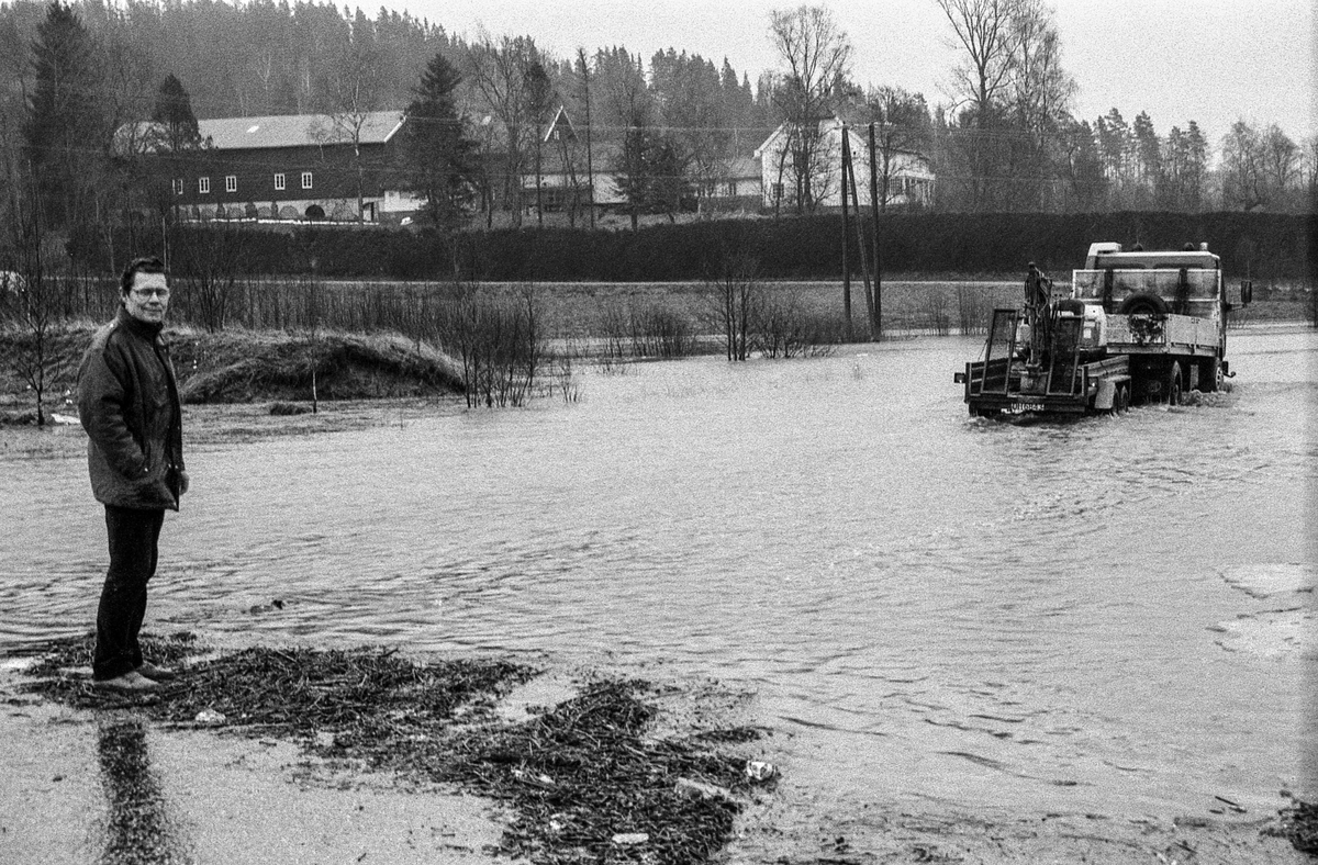 Flomskader på veiene, Riksvei 120, Ytre Enebakk. Vannet flommer over veier og jorder, biler kjører så vannspruten står.