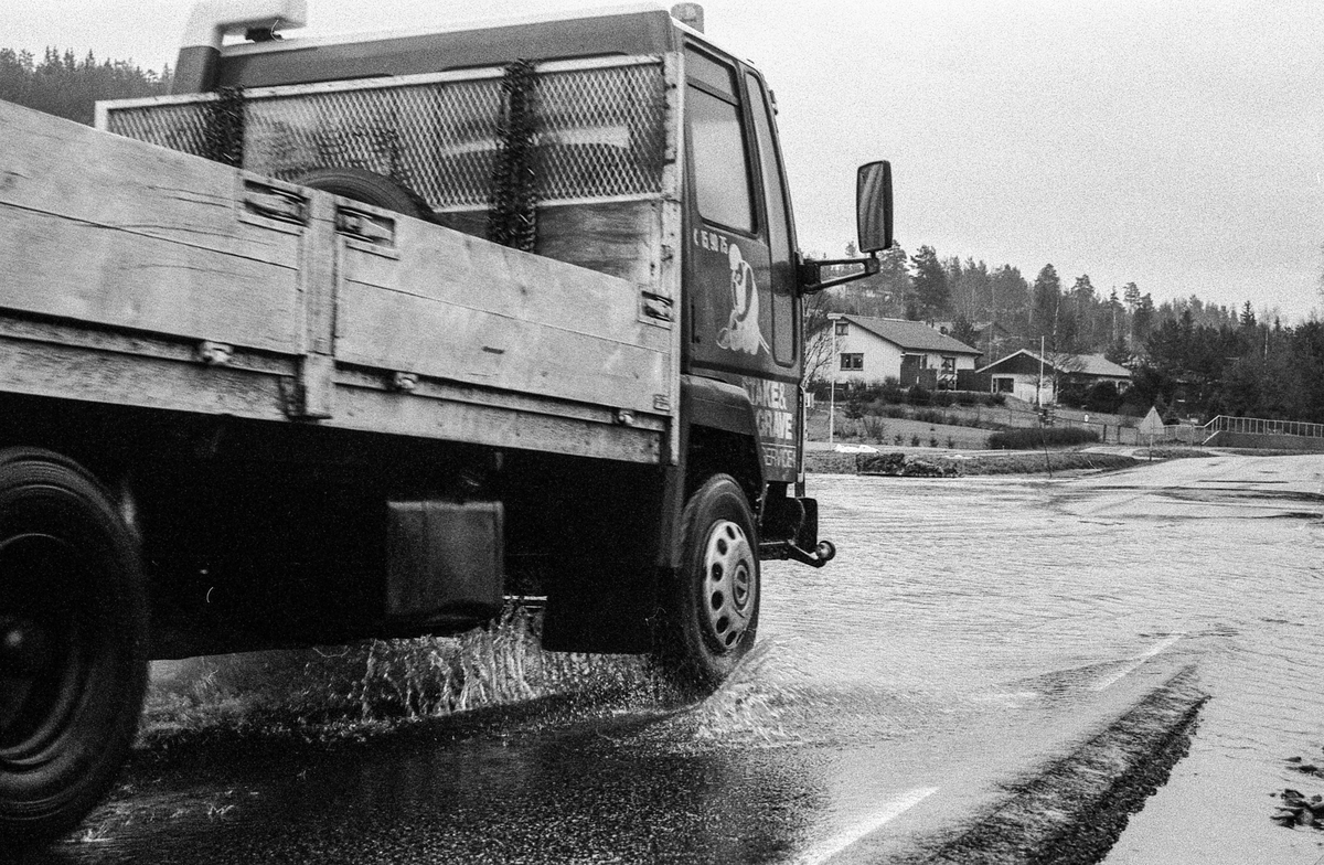 Flomskader på veiene, Riksvei 120, Ytre Enebakk. Vannet flommer over veier og jorder, biler kjører så vannspruten står.
