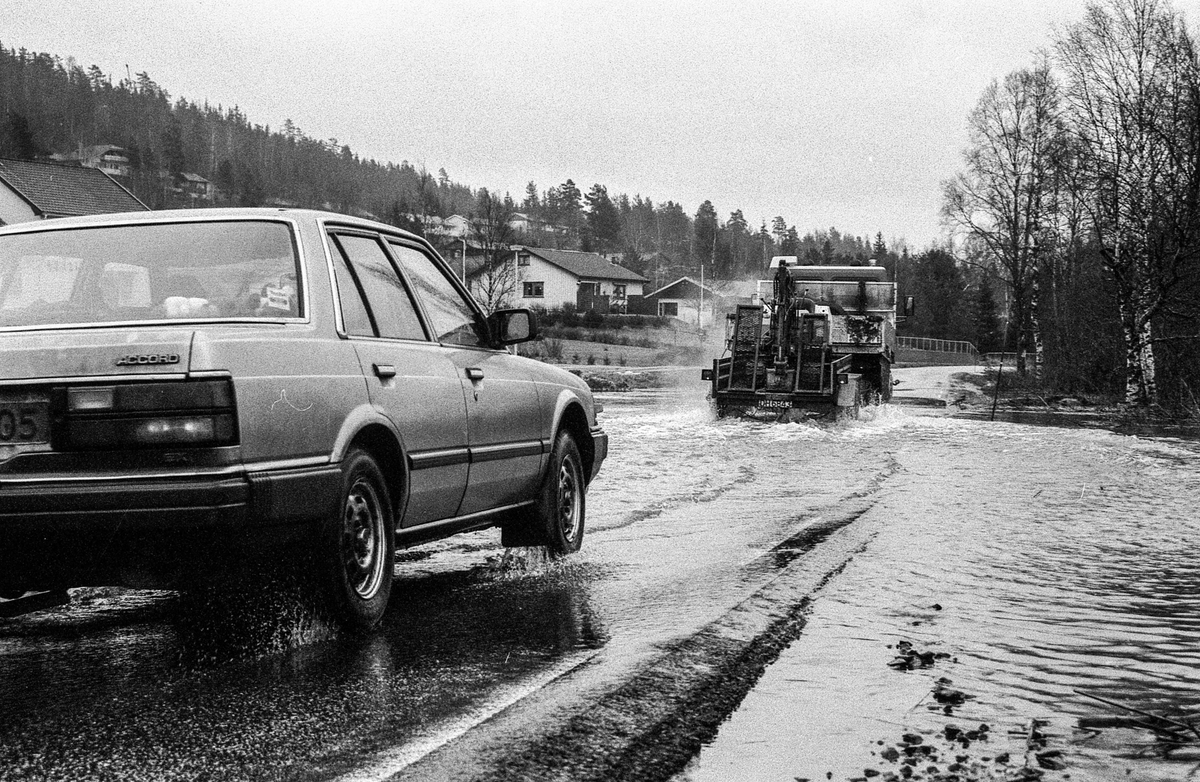 Flomskader på veiene, Riksvei 120, Ytre Enebakk. Vannet flommer over veier og jorder, biler kjører så vannspruten står.