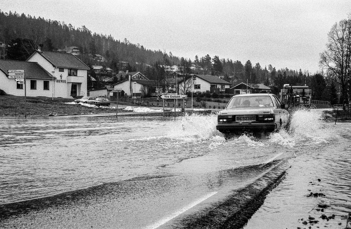 Flomskader på veiene, Riksvei 120, Ytre Enebakk. Vannet flommer over veier og jorder, biler kjører så vannspruten står.