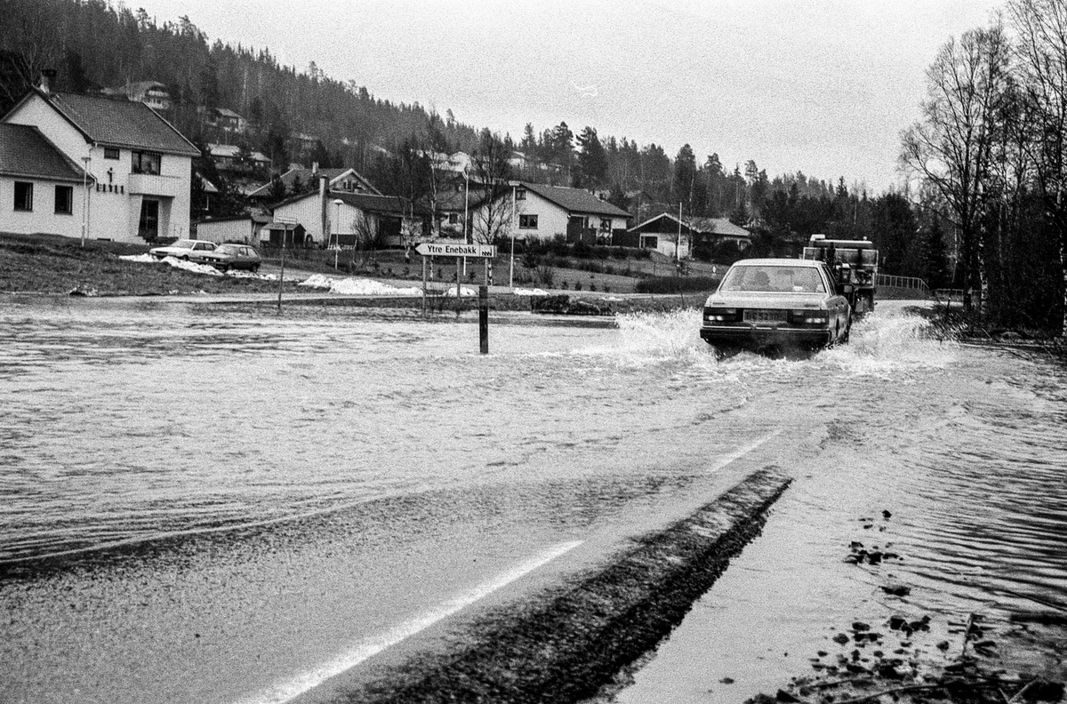 Flomskader på veiene, Riksvei 120, Ytre Enebakk. Vannet flommer over veier og jorder, biler kjører så vannspruten står.