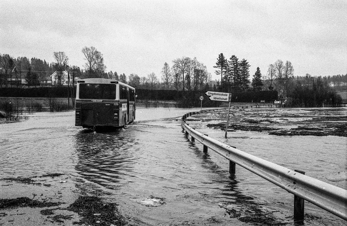 Flomskader på veiene, Riksvei 120, Ytre Enebakk. Vannet flommer over veier og jorder, biler kjører så vannspruten står.