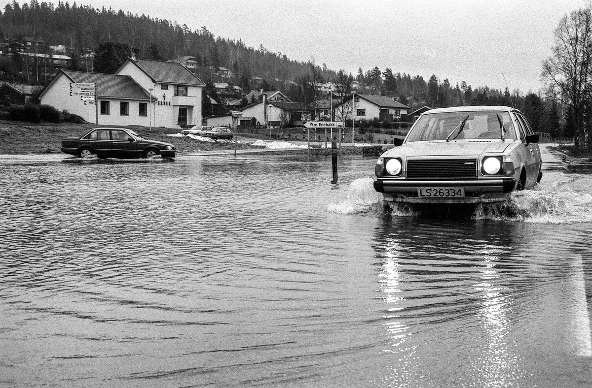 Flomskader på veiene, Riksvei 120, Ytre Enebakk. Vannet flommer over veier og jorder, biler kjører så vannspruten står.
